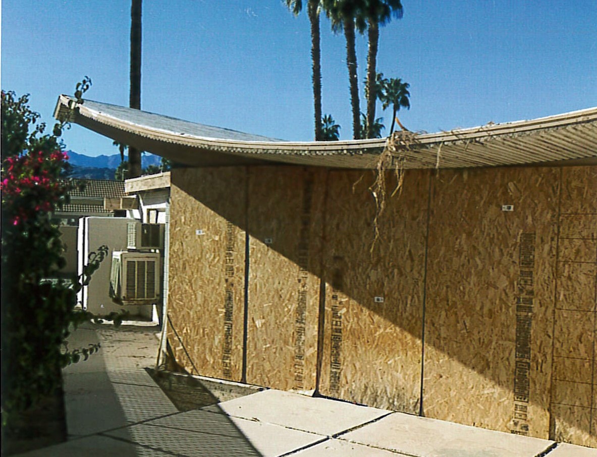 View of southwest corner of house, windows are boarded up, view additions in background